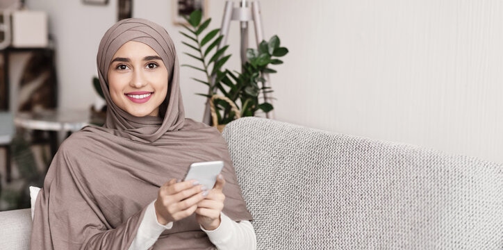 Portrait of smiling muslim woman in hijab using smartphone at home, relaxing on couch and looking at camera, free space