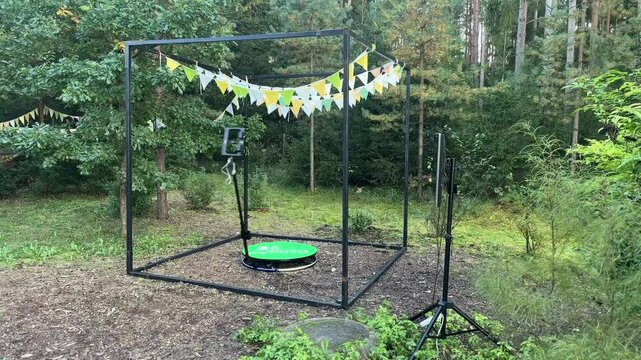 Valmiera, Latvia - September 19, 2024 -  Outdoor 360-degree photo booth setup with flags in a forested area, featuring a rotating platform and camera stand for event photography.