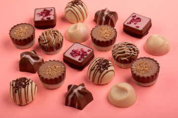photo shows an assortment of assorted chocolates placed on a pink background. The chocolates come in various shapes, sizes, and designs, some decorated with nuts, drizzles, or other toppings