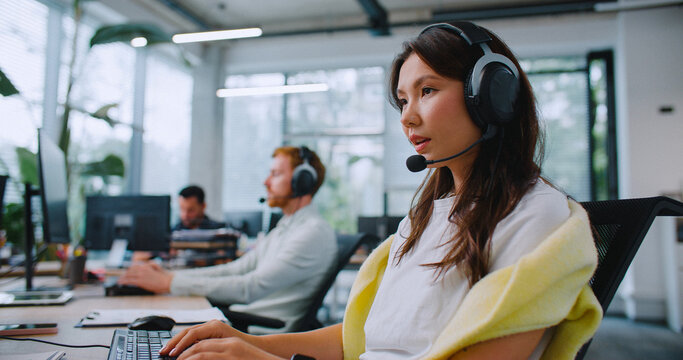Side view of hardworking operators using headsets while consulting customers from office. Workers sitting at computer and entering information on keyboard. Providing support for clients.