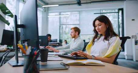 Beautiful woman actively typing text on keyboard and looking at display. Managers working at their computers in background. Office routine.