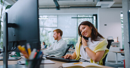Satisfied woman speaking with someone on smartphone in office. Working at computer and typing text...