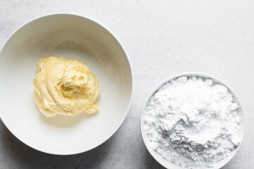 Overhead view of butter and icing sugar in a white mixing bowl, top view of margerine and powdered sugar on a marble countertop, ingredients for making buttercream, making american buttercream
