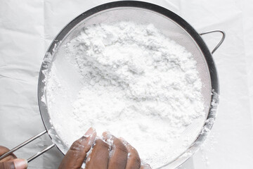 Overhead view of icing sugar in a silver sieve, sifting powdered sugar on a white table, top view of powdered sugar being sieved on a marble countertop