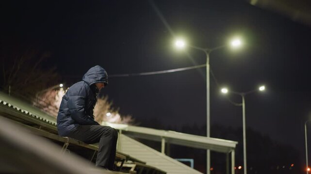 A man wearing a hood is seated alone on bleachers at night, with streetlights illuminating the surrounding area