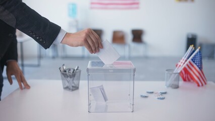 Fototapeta premium Side view of man throwing ballot into voting basket at polling station. Voter making choice while electing for desired candidate for president. American flags hanging on wall. Nation of patriots.