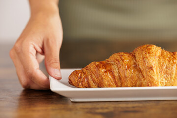 Person serving handmade croissant in bakery. Close-up portrait of croissant on white plate. The perfect breakfast, grilled croissant