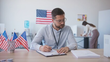 Caucasian man calmly sitting at desk and filling in information on piece of paper. Miniature flags...