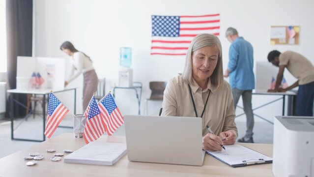 American citizens visiting local polling station. Smiling woman meeting voters while sitting at table with laptop. Poll worker checking documents and giving voting forms. Choosing president.