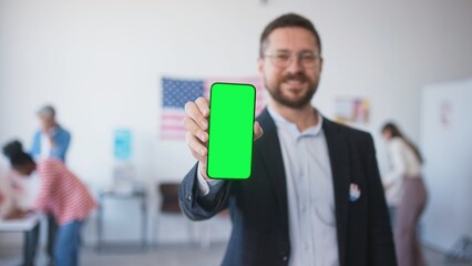 Camera focus on man's hand holding cell phone with green screen. Blurred male showing green display. People filling out ballots while choosing new candidate for President in polling station.