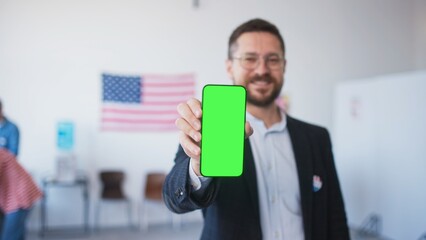 Camera focus on man's hand holding cell phone with green screen. Blurred male showing green display. People filling out ballots while choosing new candidate for President in polling station.