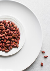 Overhead view of unpeeled raw peanuts on a white plate, flatlay of raw peanuts on a marble countertop, process of making roasted peanuts