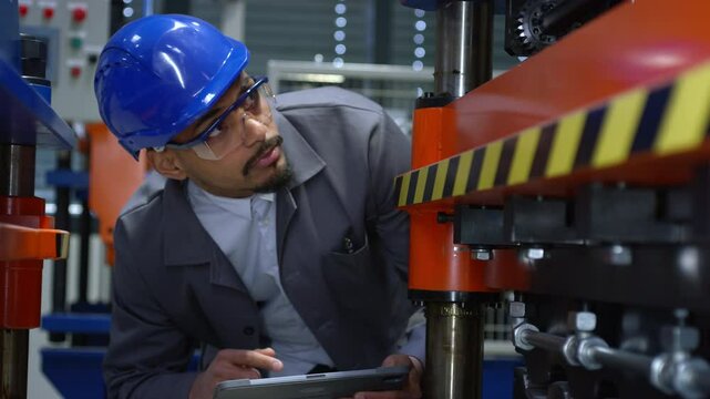 Engineer, with a blue helmet and safety glasses, checking manufacturing machinery, regulating the automatization process on a tablet, close up shot.