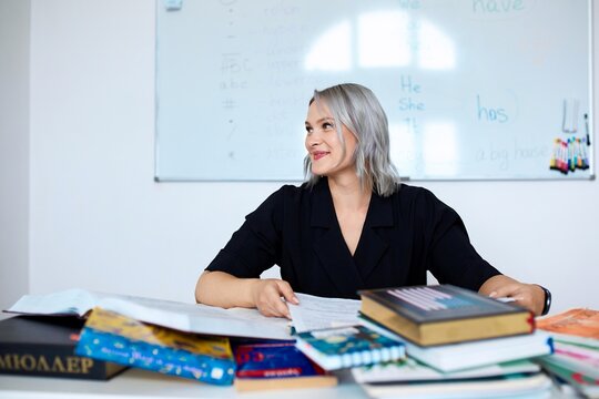Portrait of a young beautiful English teacher working at school, at her desk with a bunch of books.