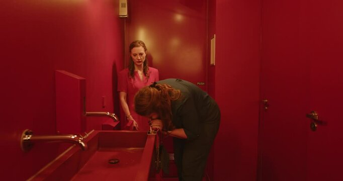 Two woman doing drugs in a public bathroom. One woman instructs the other in how to snort cocaine from the mirror