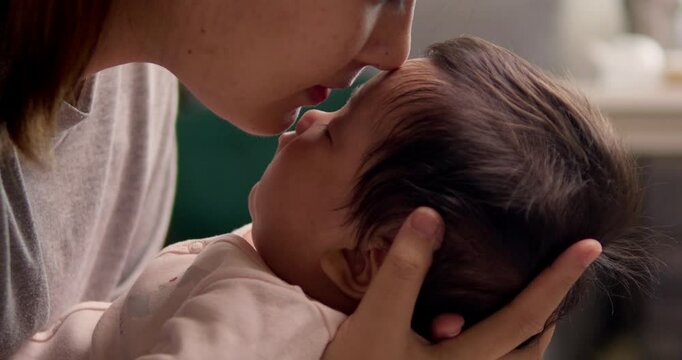 A mother leans down to smell her newborn child's head and gently kisses her baby on the forehead.