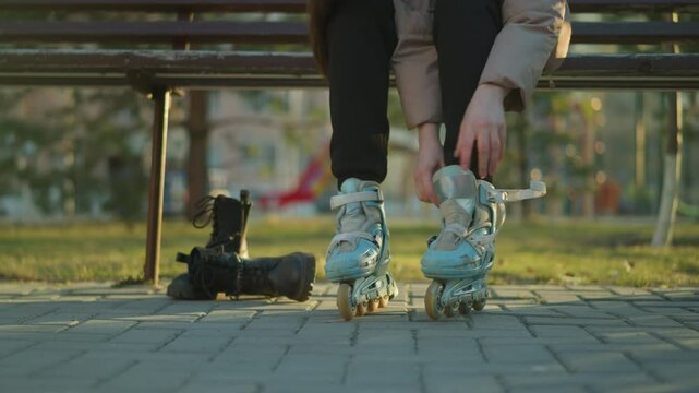 A close-up shot of a person sitting on a park bench, fastening the straps of a rollerblade on one foot while the other foot is already in a rollerblade. The person is not fully visible