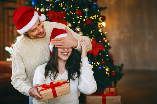 Loving young handsome man giving surprise gift to his wife, closing her eyes, celebrating Christmas at home