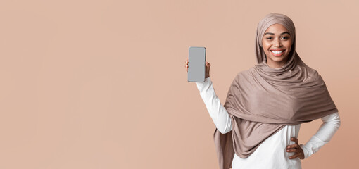 Happy afro muslim woman in hijab demonstrating modern smartphone with blank screen for mockup, posing over yellow background, free space