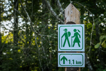 A hiking trail sign features green silhouettes of a man and woman carrying walking sticks. The sign is mounted on a weathered wooden post, set against a blurred natural background.