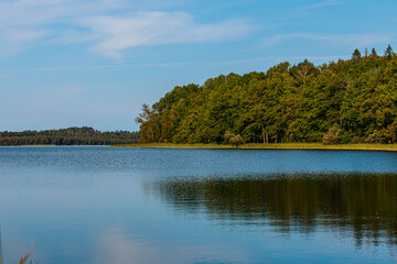 A serene view of Augstroze Lake, with calm blue waters in the foreground and a tree-lined shore in the distance. The trees exhibit early autumn colors, under a clear sky.