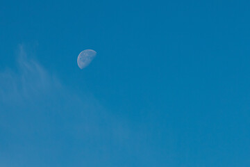 A high-resolution image of the moon in its first quarter phase, set against a clear blue sky with faint wisps of clouds. Minimalist composition with copy space.