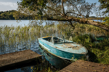 Obraz premium An old, weathered blue boat rests near a wooden dock at the edge of a serene lake, surrounded by tall reeds and overhanging tree branches. The calm water and nature create a peaceful