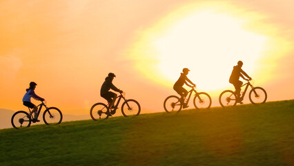 AERIAL, SILHOUETTE, LENS FLARE: Stunning sunset with group of mountain bikers riding up the grassy hill. Picturesque bike ride through the green, hilly Slovenian countryside in beautiful golden light.