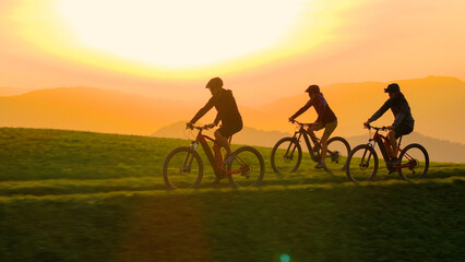 AERIAL, SILHOUETTE, LENS FLARE Three cyclists against a vibrant sunset, riding along a grassy hilltop as sky glows with orange and golden light. Beauty and tranquillity of an evening outdoor adventure