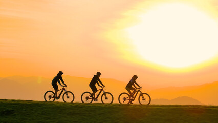 SILHOUETTE, LENS FLARE: Three mountain bikers pause on a hilltop to enjoy the serene sunset with sky ablaze in warm orange hues. Moment of connection with nature and each other after an joyful ride.