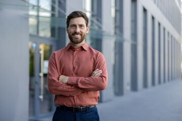 Confident male professional stands with arms crossed outside modern urban building, expressing confidence and professionalism. Businessman wearing casual attire represents successful entrepreneur