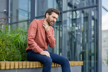 Adult male experiencing throat discomfort while seated outside in urban setting. Businessman appears concerned, capturing both health and professional stress themes.