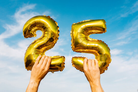 Hands holding number 25 golden balloons against blue sky