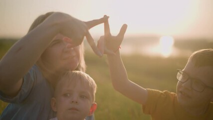 A mother in a blue dress, sitting on the grass with her two young sons during sunset. her older son in a yellow shirt, form a playful shape with their fingers while the younger boy watches intently