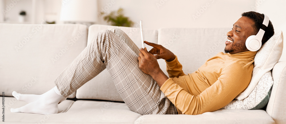 Canvas Prints Gadget Lifestyle. Positive Afro Man In Earphones Using Tablet Computer Relaxing Lying On Sofa At Home