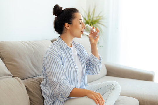 Beautiful happy calm arabian or indian brunette woman in casual wear is drinking a glass cup of clean water in the morning, while sits on sofa in living room at home, leads healthy life style concept
