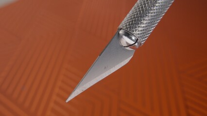 A close-up of a sharp precision craft knife blade positioned against an orange textured background. The metallic handle features a grip, ideal for detailed cutting tasks, art or DIY projects.