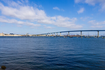 Fototapeta premium San Diego bay and the Coronado Bridge with ships sailing on blue ocean water, office buildings and skyscrapers along the coast at Coronado Tidelands Park in Coronado California