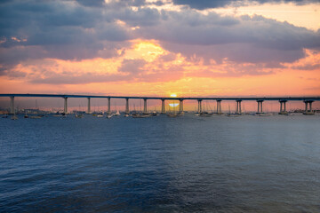 Fototapeta premium San Diego bay and the Coronado Bridge with ships sailing on blue ocean water, office buildings and skyscrapers along the coast at sunset at Coronado Tidelands Park in Coronado California