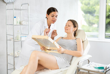Young female doctor cosmetologist analyzes face of adult female patient in medical office