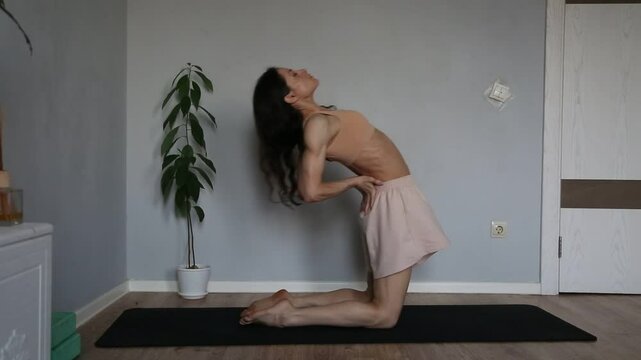 Beautiful fit atletic body woman stratching for shoulders, scapula and healthy back in bra and sporty wear doing asana camel pose on yoga mat at home interior with green flower. Ustrasana. 