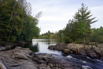 Transition between calm water and rapids on a forest-edged river with exposed rocks in the foreground