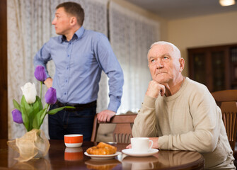 Frustrated aged man sitting separately at home table having problems in relationship with his adult son