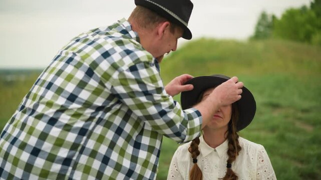 An artist in a checked shirt adjusts the hat of a woman with braided hair, wearing a white dress, seated in a green field, smiling shyly