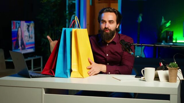 Upbeat influencer holding colorful shopping bags in hand, showing subscribers purchases he recently got. Cheerful content creator presenting fanbase new acquisitions, camera A