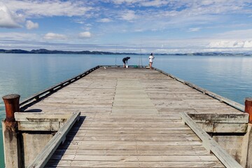 Obraz premium People fishing off a pier over a calm ocean beach in Maraetai, Auckland, New Zealand.