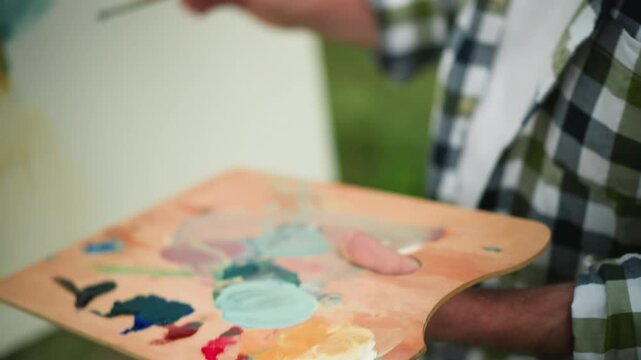 A painter delicately holds a wooden palette filled with vibrant paint as they apply final touches to their artwork. focusing on the hand and brush in action
