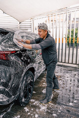 A latin man boldly washing a black car with soap foam and a sponge in the side of a car wash.