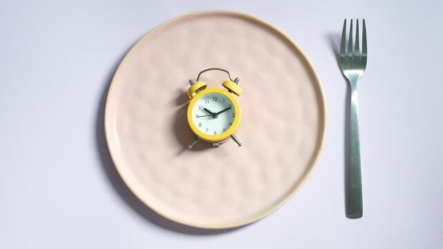 Empty plate with yellow alarm clock on lilac background top view close-up.