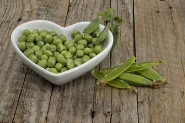 Fresh green peas in a heart-shaped bowl on a rustic wooden table, with pea pods next to the bowl